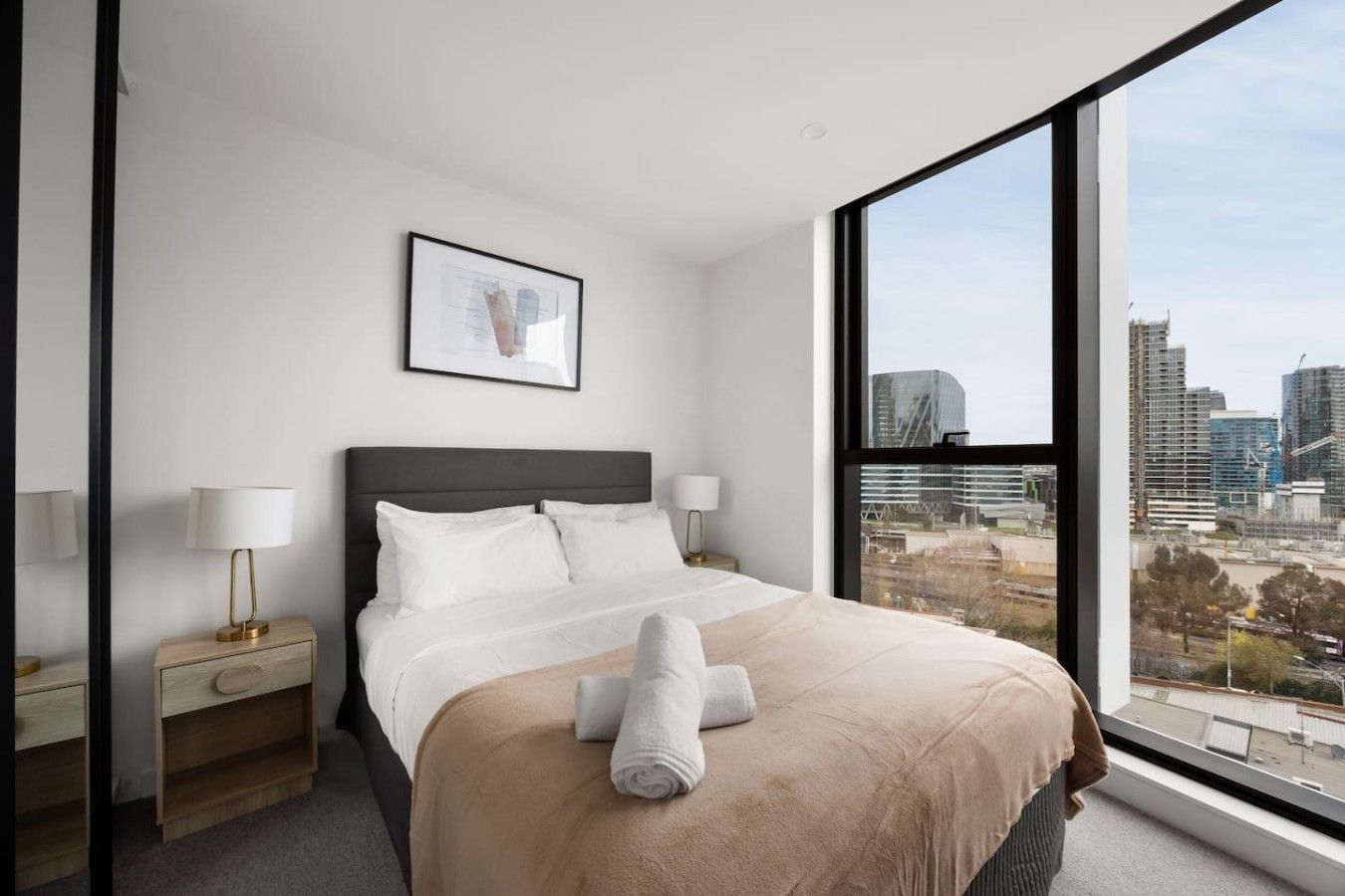 Une chambre d’hôtel moderne avec une grande fenêtre, un lit avec du linge de maison blanc, une table de chevet et un tableau encadré au mur.