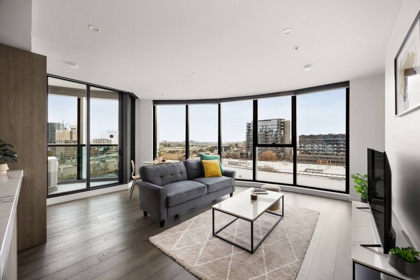A modern living room with a grey sofa, a coffee table, and large windows offering a city view.