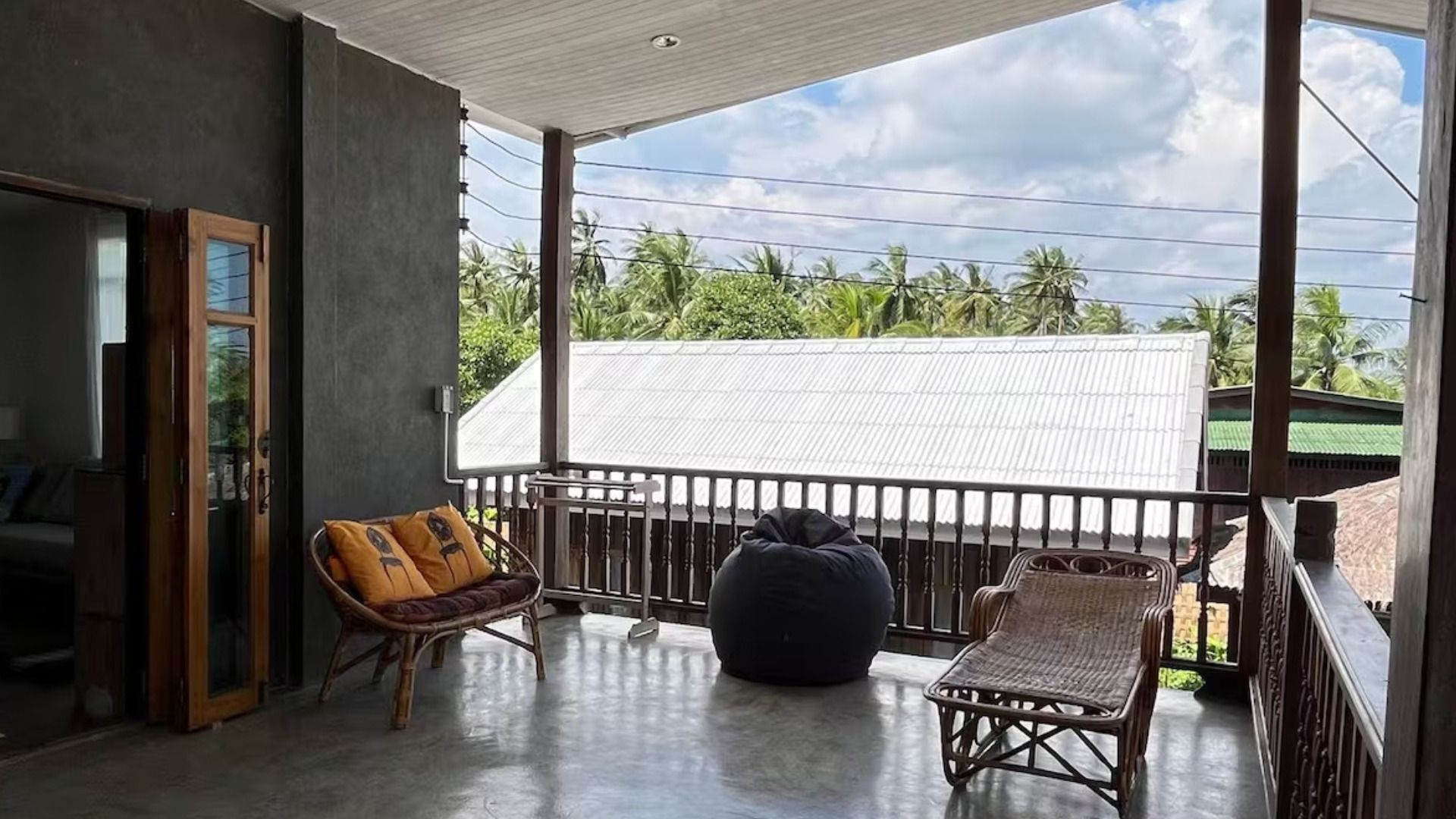 Un balcon avec vue sur des palmiers et un bâtiment blanc. On y trouve deux fauteuils en osier et un pouf noir.