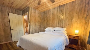 Una camera da letto con pavimento in legno, un letto con biancheria bianca, un comodino con una lampada e una porta che dà su un'altra stanza.