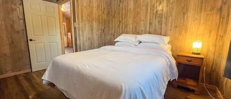 A bedroom with a wooden floor, a bed with white bedding, a nightstand with a lamp, and a door leading to another room.