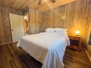 Ein Schlafzimmer mit Holzboden, einem Bett mit weißer Bettwäsche, einem Nachttisch mit Lampe und einer Tür zu einem anderen Raum.