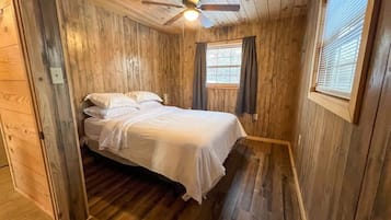 Ein Schlafzimmer einer Holzhütte mit einem Bett, einem Deckenventilator und einem Fenster mit Vorhängen.