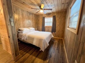 Ein Schlafzimmer einer Holzhütte mit einem Bett, einem Deckenventilator und einem Fenster mit Vorhängen.