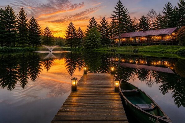 A wooden dock extending into a calm lake with a reflection of trees and a building with lights.