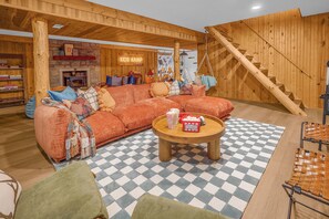 A cozy living room with a checkered floor, a wooden staircase, and a children's play area labeled 'KIDS KAMP' in the background.
