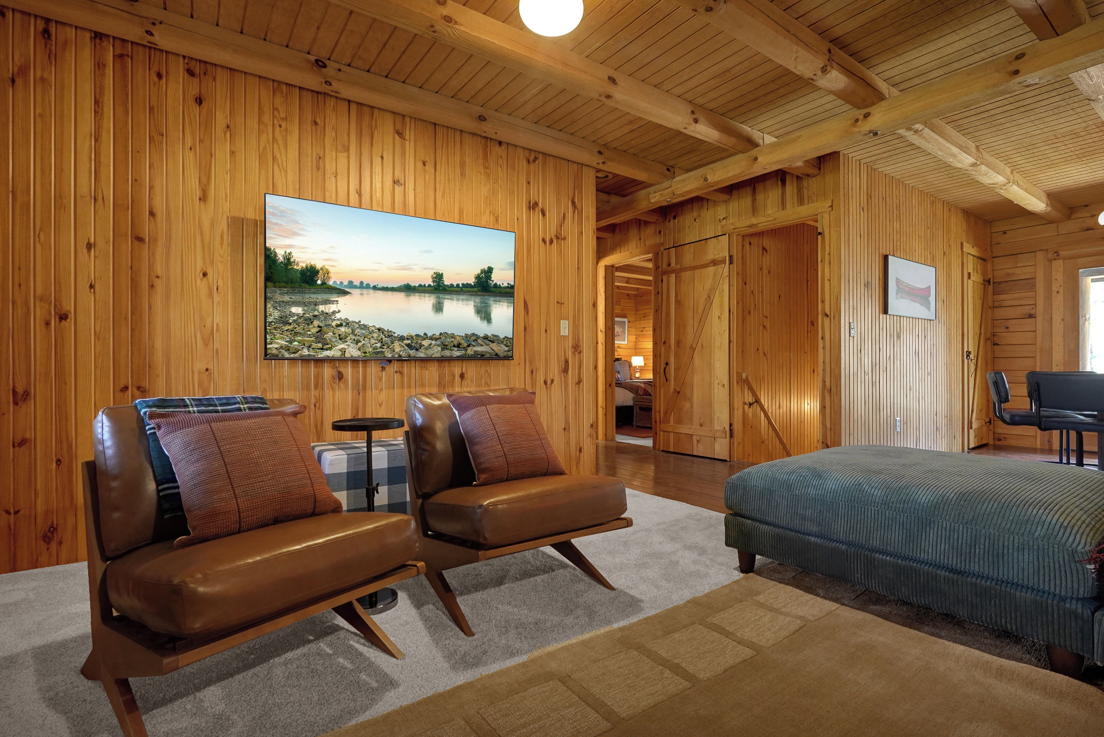 Ein Holzinnenraum mit einem Bett, zwei Ledersesseln, einem Fernseher und Blick auf einen See.