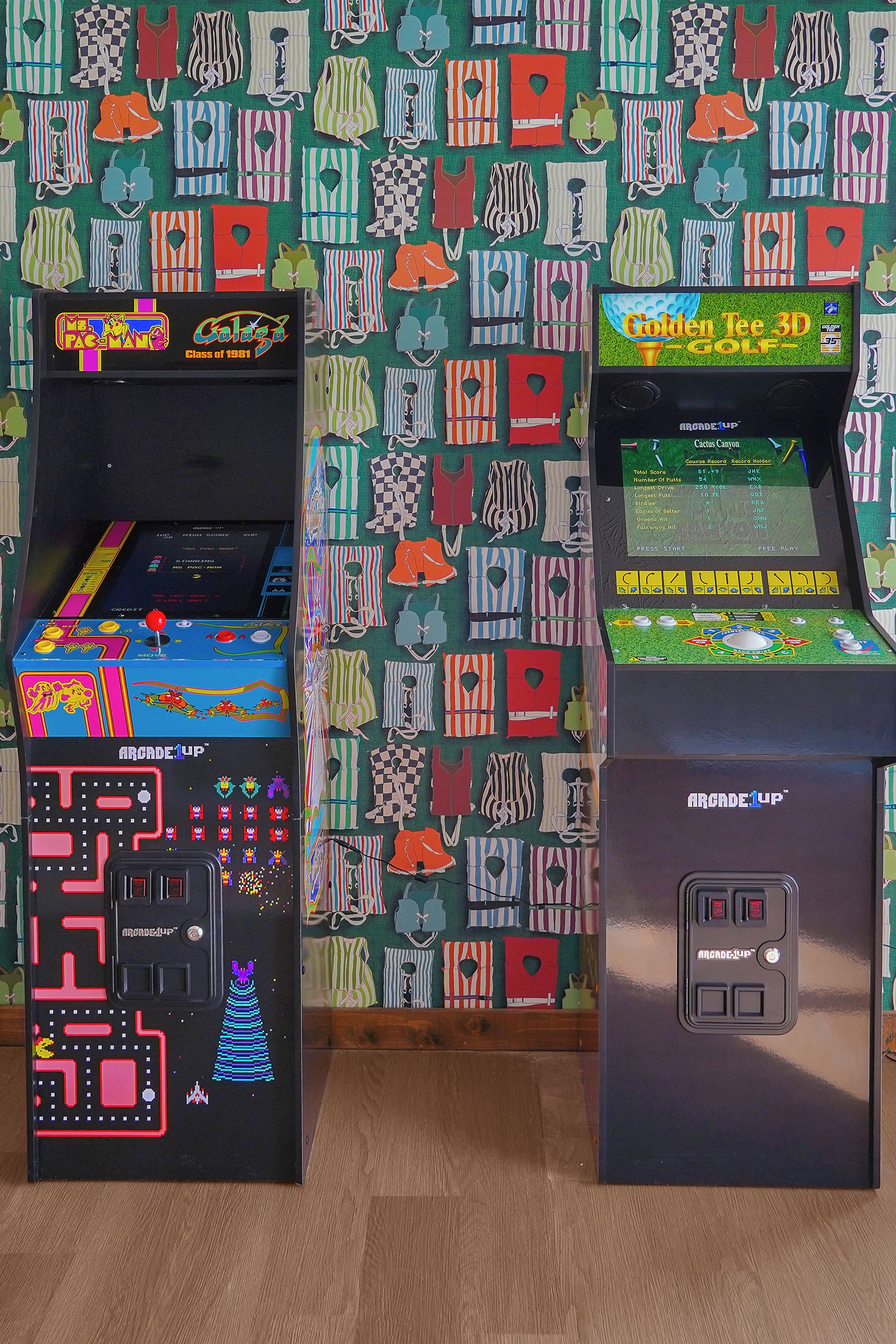 To arkademaskiner, en med Pac-Man tema og den anden med golf tema, placeret foran en væg med tapet.