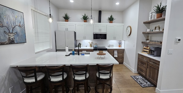 A modern kitchen with a central island, bar stools, white cabinetry, and a coffee bar.