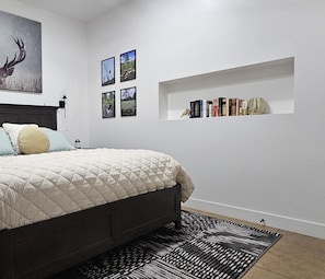 A bedroom with a bed, a wall-mounted shelf with books, and framed pictures.