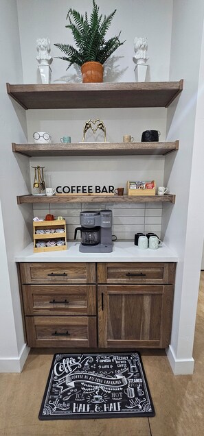 A kitchen area with a coffee bar, shelves with decorative items, a coffee machine, and a coffee-themed rug.