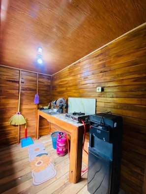 A room with wooden walls, a desk with a computer, a fan, a lamp, and a pink fire extinguisher.