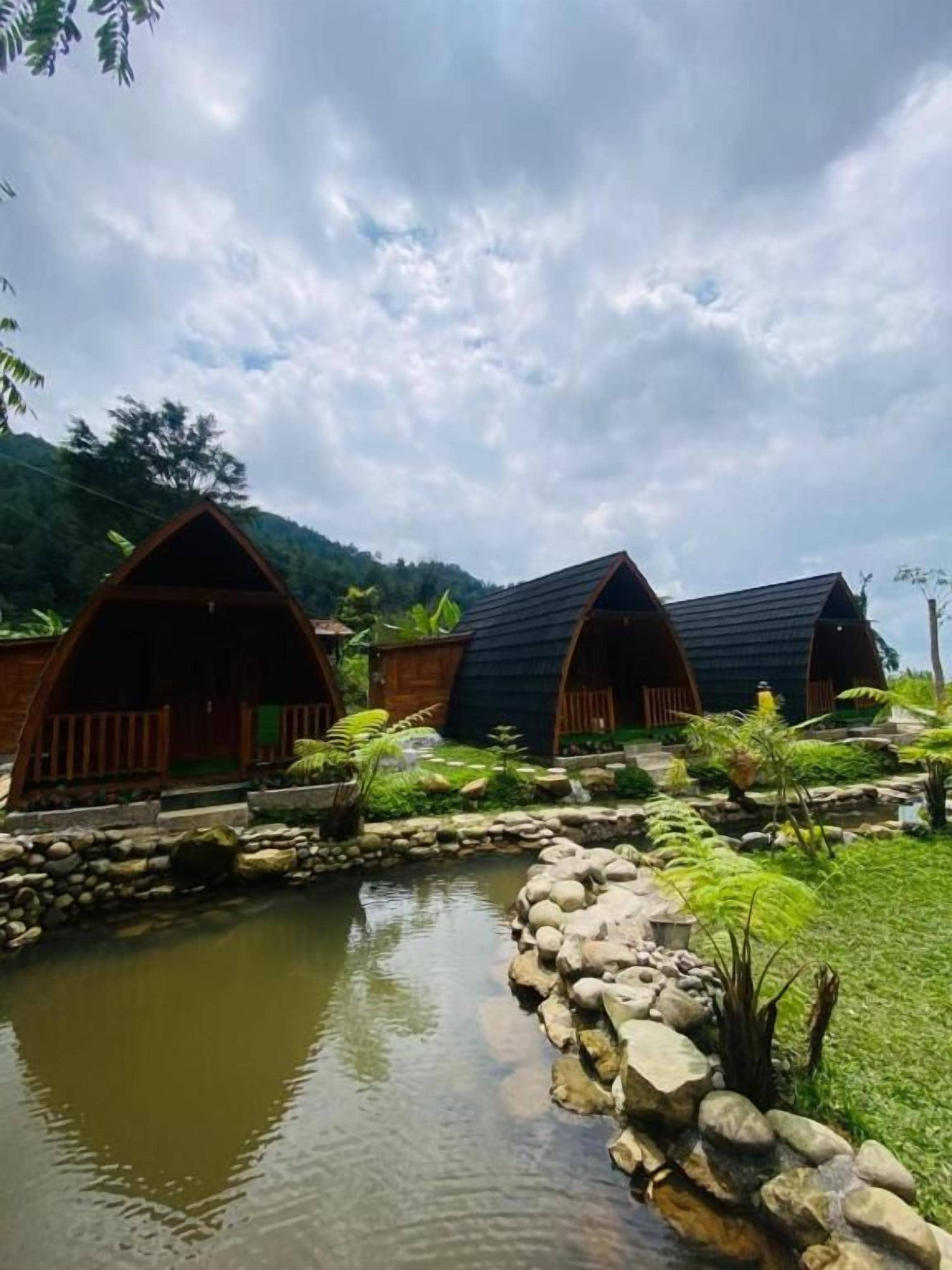 A row of wooden cabins with sloped roofs, a small pond, and a rocky path leading to them.