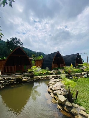A row of wooden cabins with sloped roofs, a small pond, and a rocky path leading to them.