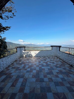 Una terraza con paredes de piedra, suelo de baldosas y barandilla de metal, con vistas a un paisaje montañoso.