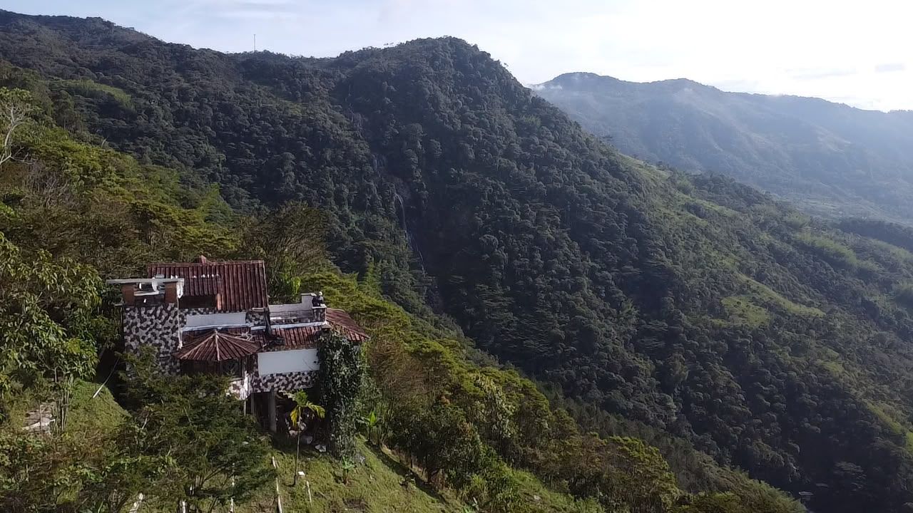 Paisaje montañoso con una pequeña construcción de techo rojo y paredes blancas, rodeada de abundante vegetación.