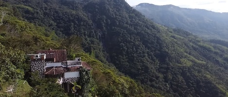 A mountainous landscape with a small building featuring a red roof and white walls, surrounded by dense greenery.