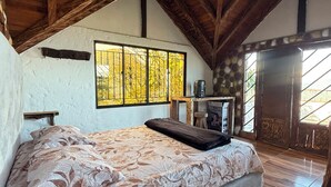 Quarto com cama grande, vigas de madeira no teto, janela com vista e uma pequena mesa com cadeira.