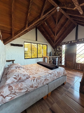 A bedroom with a large bed, wooden ceiling beams, a window with a view, and a small table with a chair. - El Colibrí Cabin in Cocorná: Incredible View (Cocorná)