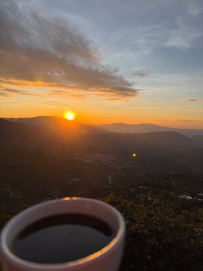 Un paysage avec une tasse de café au premier plan, un coucher de soleil en arrière-plan et une vallée entourée de montagnes.