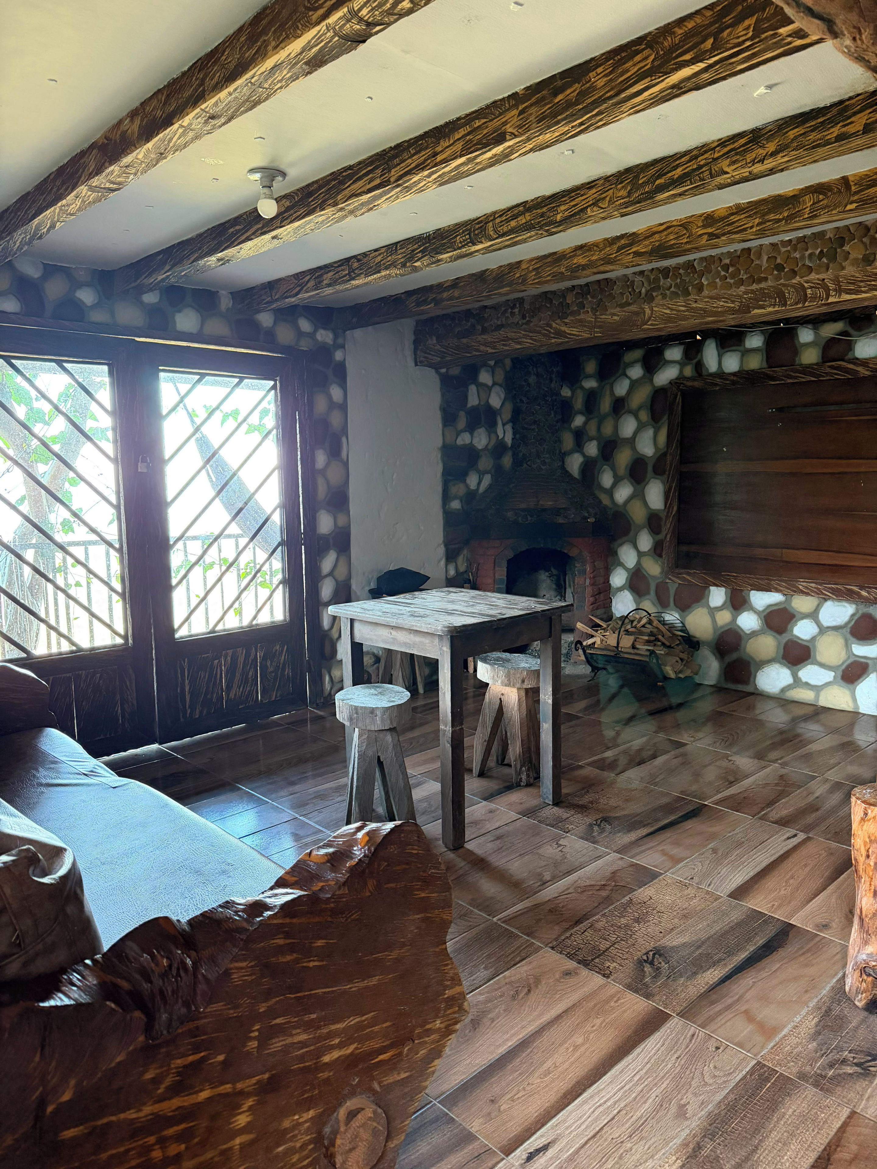 Una habitación rústica con pared de piedra, vigas de madera, chimenea y una mesa con taburetes.