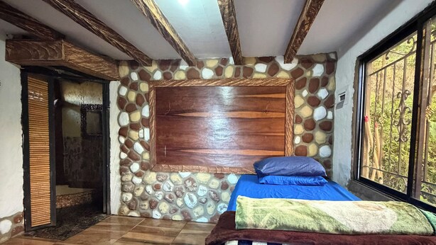 Quarto com parede de pedra, teto de madeira, cama com colcha azul e janela com grades.