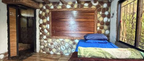 A bedroom with a stone wall, wooden beams, a bed with blue bedding, and a window with a view.