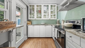 A kitchen with white cabinets, a built-in oven, a sink, and a window overlooking a garden. - StayHaus - The Meridian Flat (St. Petersburg)