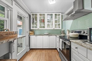 A kitchen with white cabinets, a built-in oven, a sink, and a window overlooking a garden. - StayHaus - The Meridian Flat (St. Petersburg)
