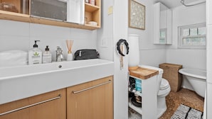 A bathroom with a white sink, mirror, and a wooden vanity. There is a toilet with a wicker basket beside it, and a shower area with a white tub. - StayHaus - The Meridian Flat (St. Petersburg)