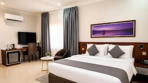 Quarto de hotel com uma cama grande, escrivaninha com televisão, cadeira e um quadro na parede.