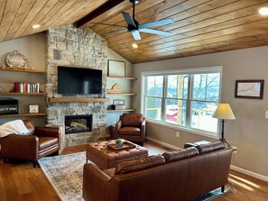 A living room with a stone fireplace, leather furniture, a flat-screen TV, and a large window with a view. - Mountain Glory - New Modern Cabin with Hot Tub and Mountain Views (Rileyville)