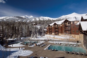 Un complexe hôtelier doté d’une piscine, entouré de montagnes enneigées, et comprenant un grand bâtiment avec de nombreux balcons.