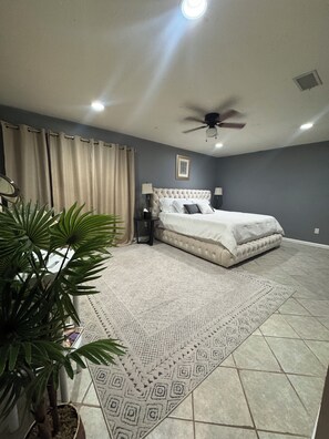 Ein Schlafzimmer mit einem Bett, Deckenventilator, Nachttisch, Lampe, Blumentopf und einem gemusterten Teppich.