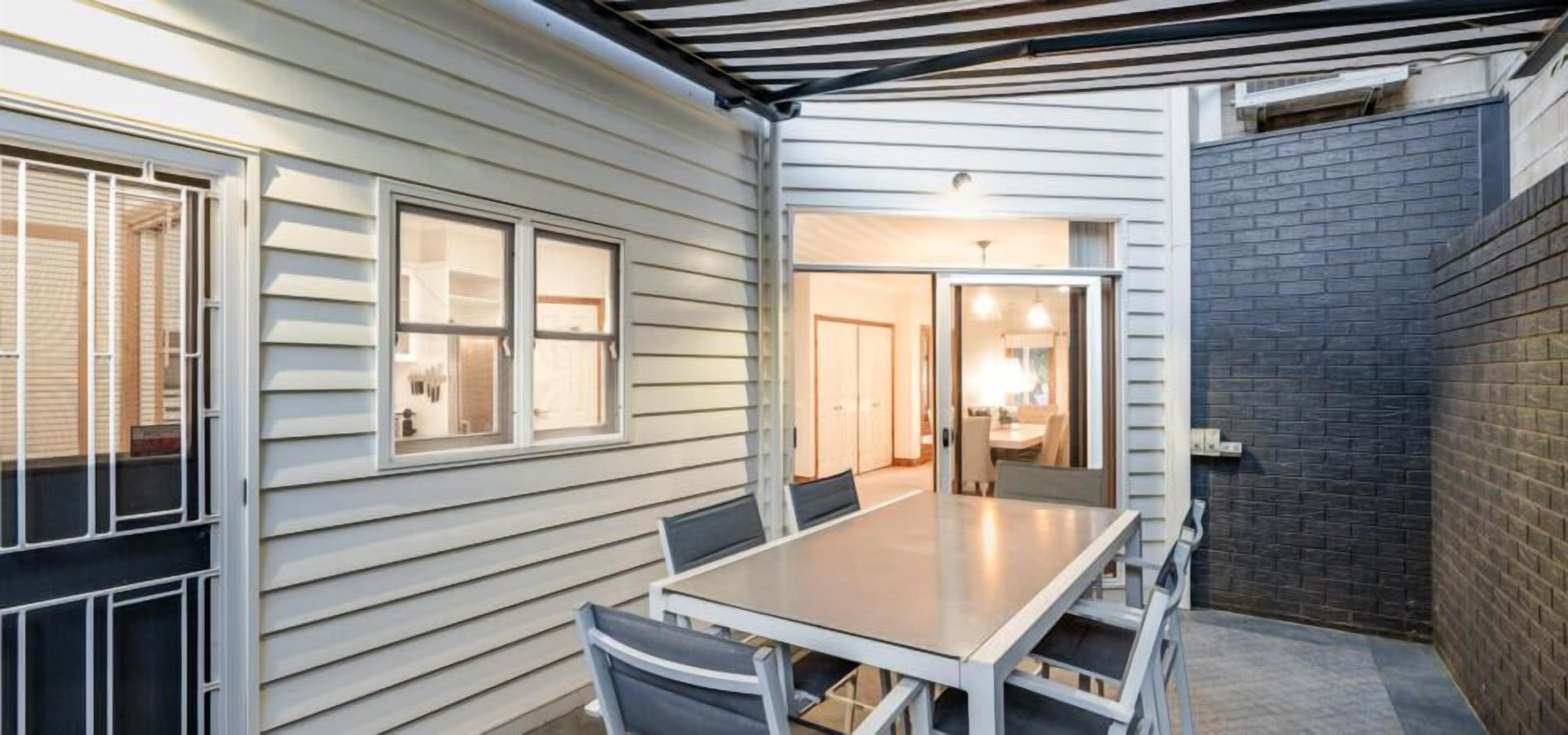 A covered patio with a dining table and chairs, a brick wall, and a door leading to an interior space.