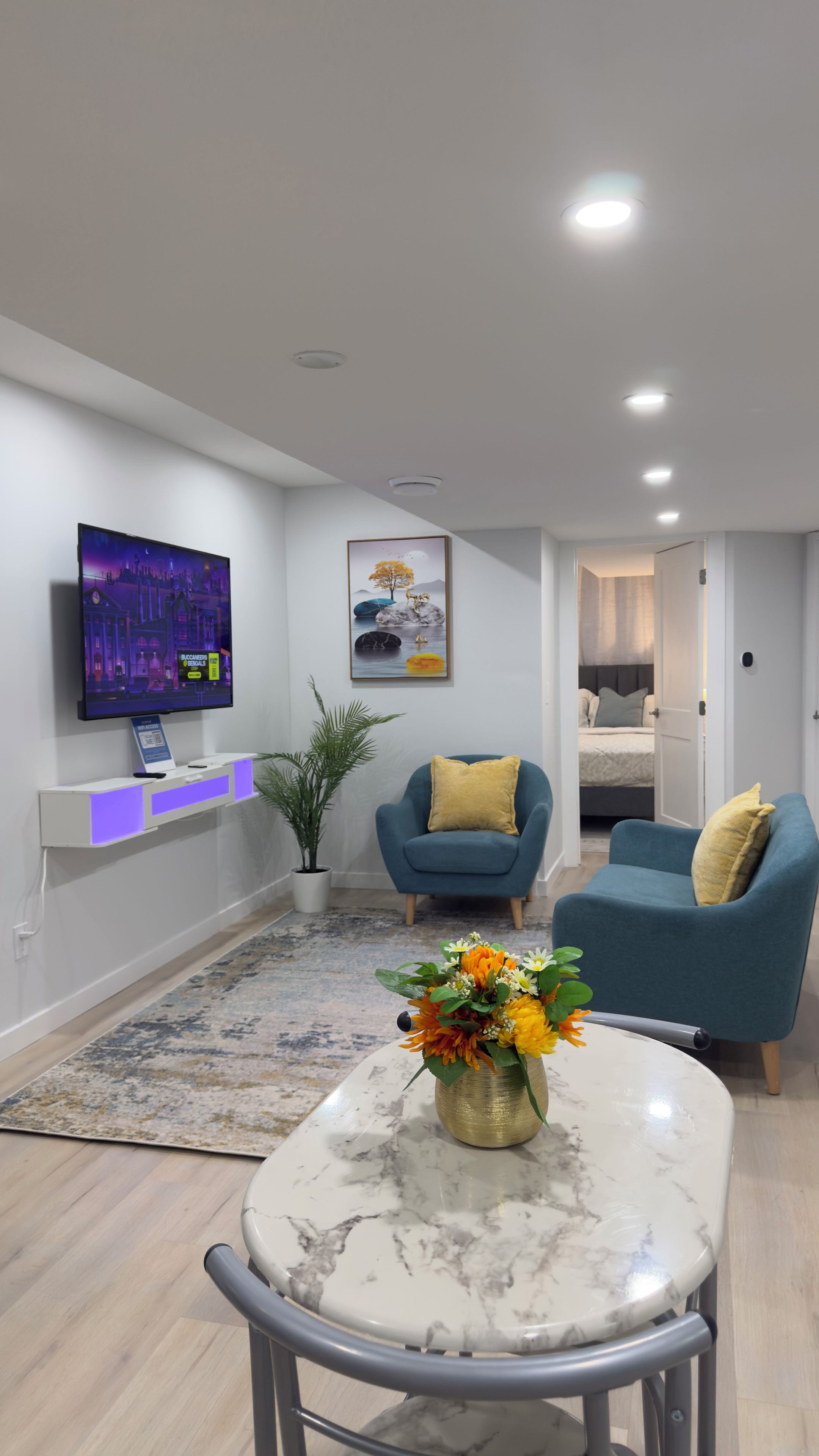 A modern living room with a flat-screen TV, a coffee table with a vase of flowers, two blue armchairs, and a bed in the background.