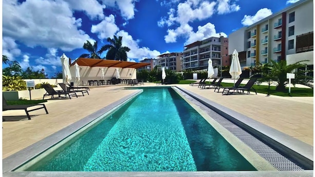 A swimming pool with lounge chairs and umbrellas, surrounded by buildings and palm trees. - Modern Maho Studio Near Mullet Bay & Maho Beach (Maho)