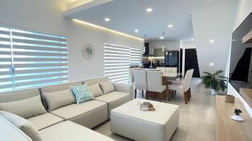 A modern living room with a sectional sofa, a dining area with a table and chairs, and a kitchen in the background.