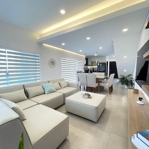 A modern living room with a sectional sofa, a dining area with a table and chairs, and a kitchen in the background.