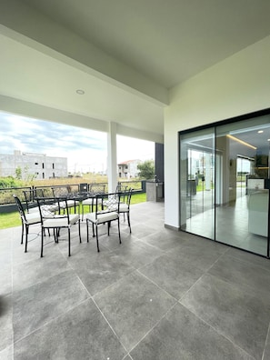 A covered patio area with a dining table and chairs, a glass door leading to an indoor space, and a view of the surrounding buildings.