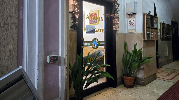 Entrada de hotel com balcão de recepção, placa 'AKIN GATE' e um letreiro de 'BEM-VINDO'.