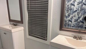 Un baño con lavamanos blanco, un espejo y una ventana con persianas.