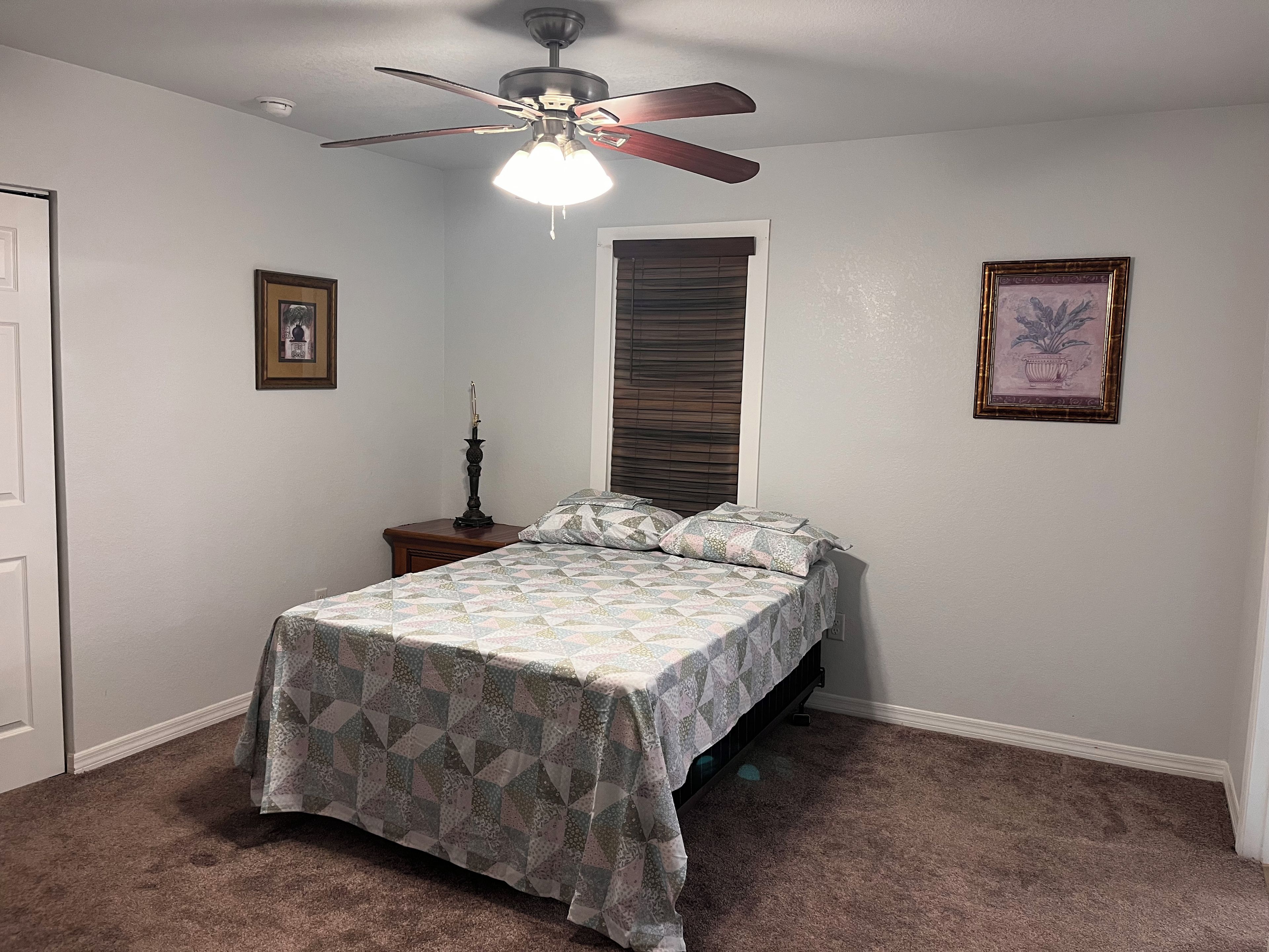 A bedroom with a bed, a nightstand, a ceiling fan, a window with blinds, and two framed pictures on the wall.