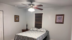 Un dormitorio con cama, mesita de noche, ventilador de techo, ventana con persianas y dos cuadros en la pared.