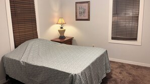 Un dormitorio con una cama, una mesita de noche con lámpara, dos ventanas con persianas y un cuadro en la pared.