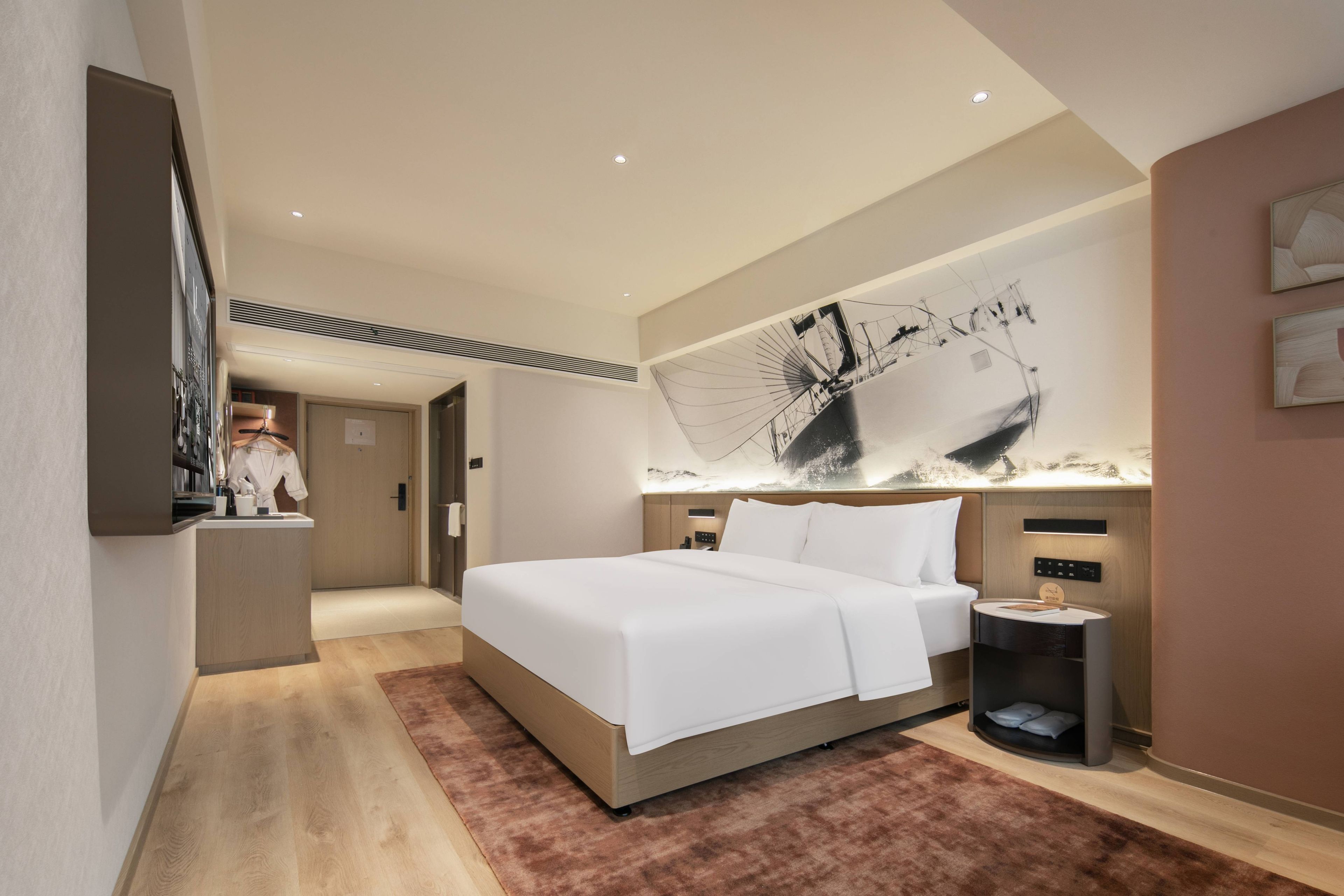 A modern hotel room with a large bed, a bedside table, a nightstand, a wall-mounted artwork, and a hallway leading to a bathroom.