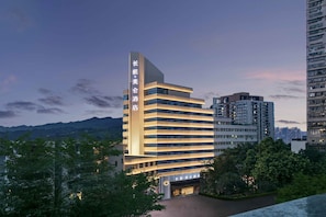 Un moderno edificio de hotel con un llamativo letrero vertical, varios pisos y una entrada bien iluminada.
