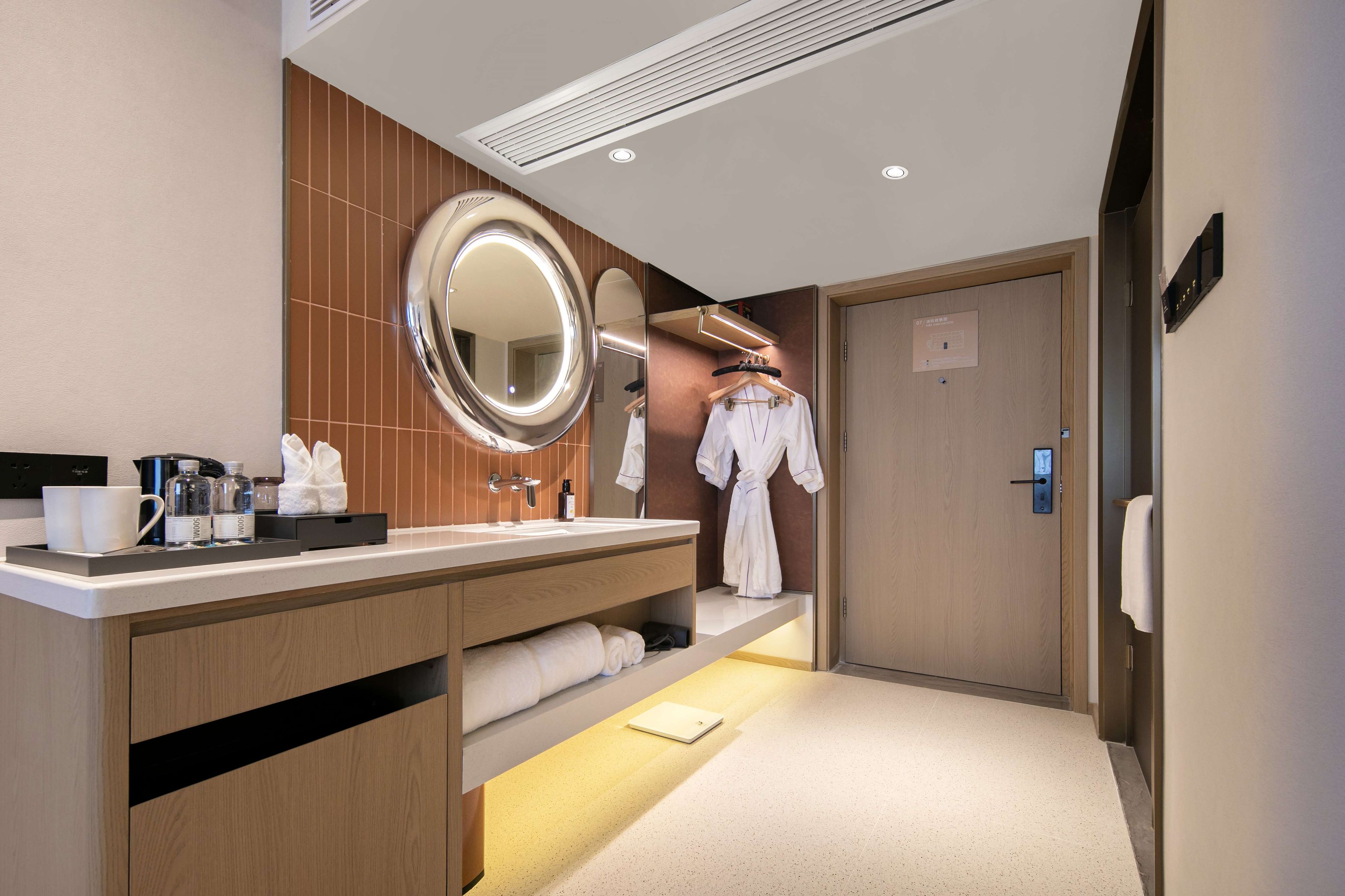 A hotel bathroom with a vanity, mirror, towel rack, and a robe hanging on the wall.