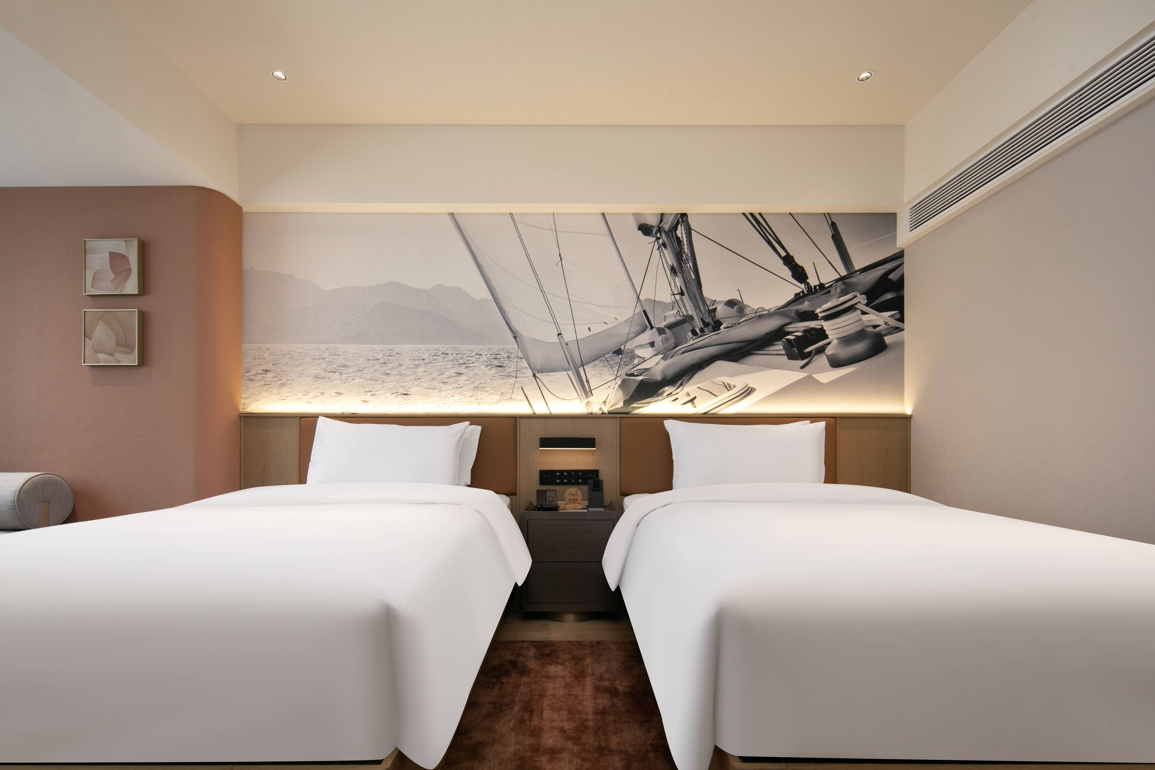 Een hotelkamer met twee bedden, een groot wandtapijt van een zeilboot en een klein nachtkastje met een lamp.