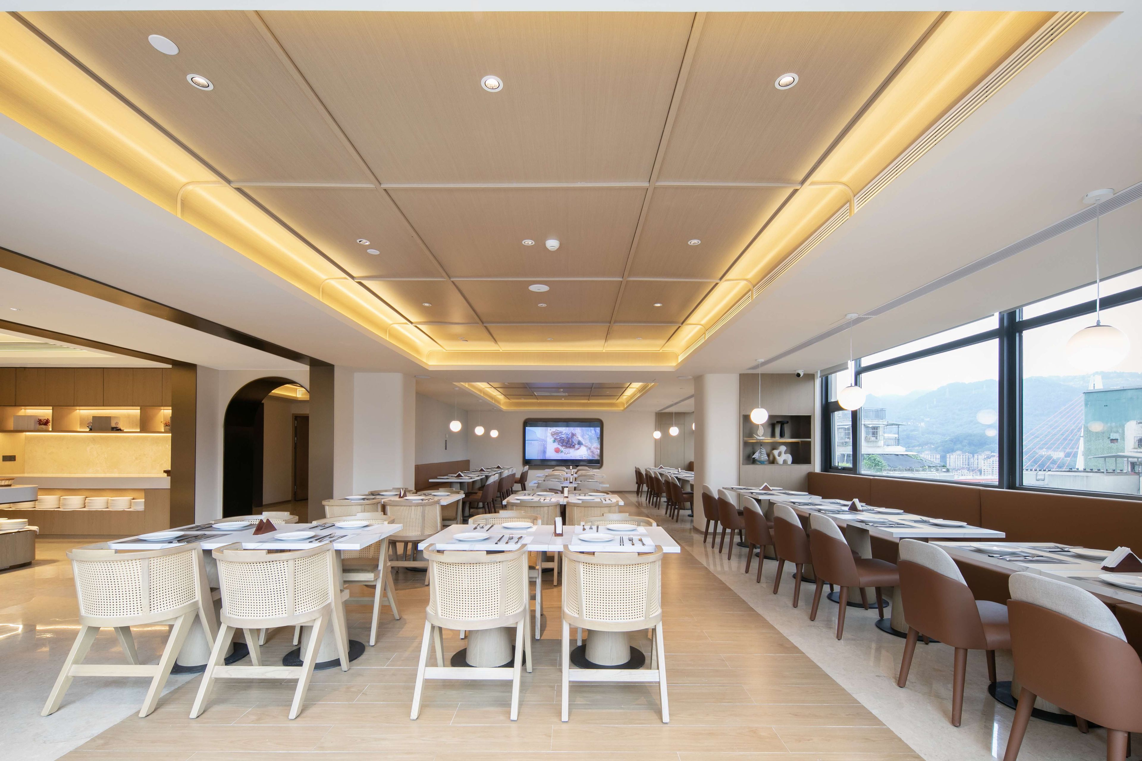A modern dining area with tables and chairs, a television screen, and a view of the city through large windows.